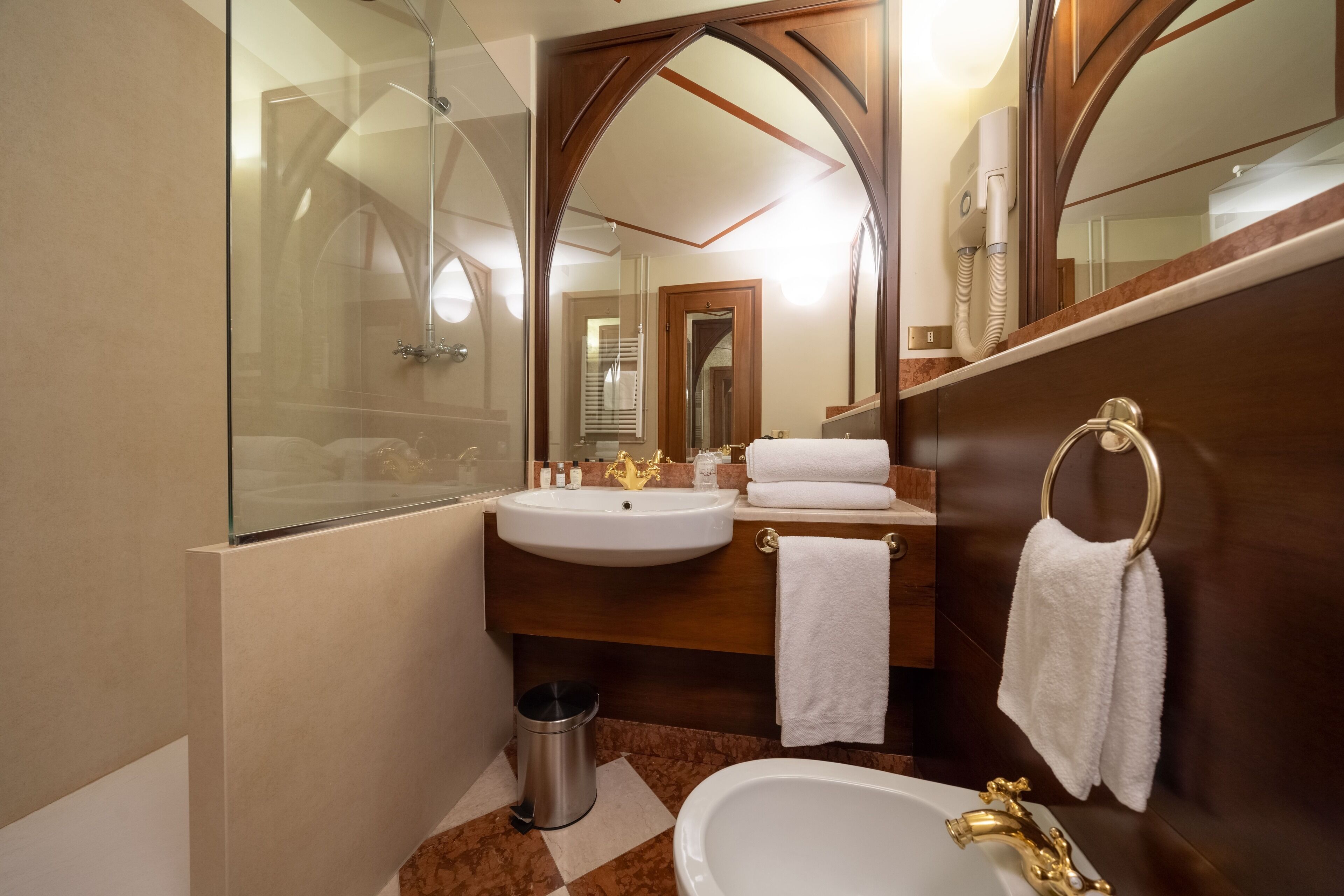 Een moderne badkamer met een glazen douchecabine, een wastafel met gouden kraan en een bad met gouden kraan. Er hangen twee handdoeken aan de muur en een prullenbak staat in de buurt van de wastafel.
