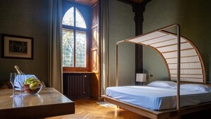 A room with a wooden floor, a bed with a curved canopy, a table with a fruit bowl, a framed picture, and a window with a view of trees.