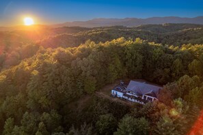 An aerial view of a house surrounded by dense forest, with a clear sky and distant mountains.