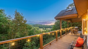 Una terraza de madera con barandilla, mobiliario exterior y vistas a un paisaje boscoso.