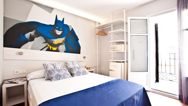 A hotel room with a large Batman poster, a neatly made bed with blue bedding, a small bedside table with a magazine, and a wardrobe with a mirror. - Ryans Pocket (Ibiza Town)