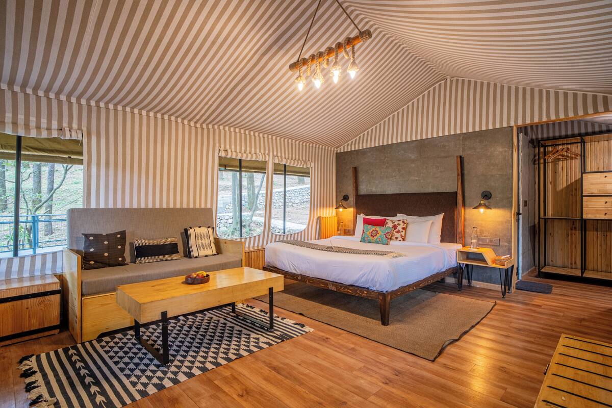 A room with a bed, sofa, and a small table. There are windows and a view of trees outside.