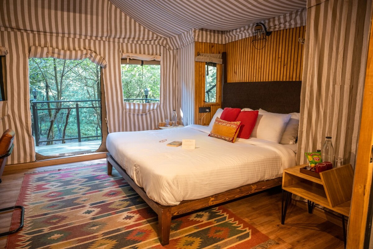 A bedroom with a large bed, a wooden headboard, striped curtains, a view of trees, and a small table with a chair.