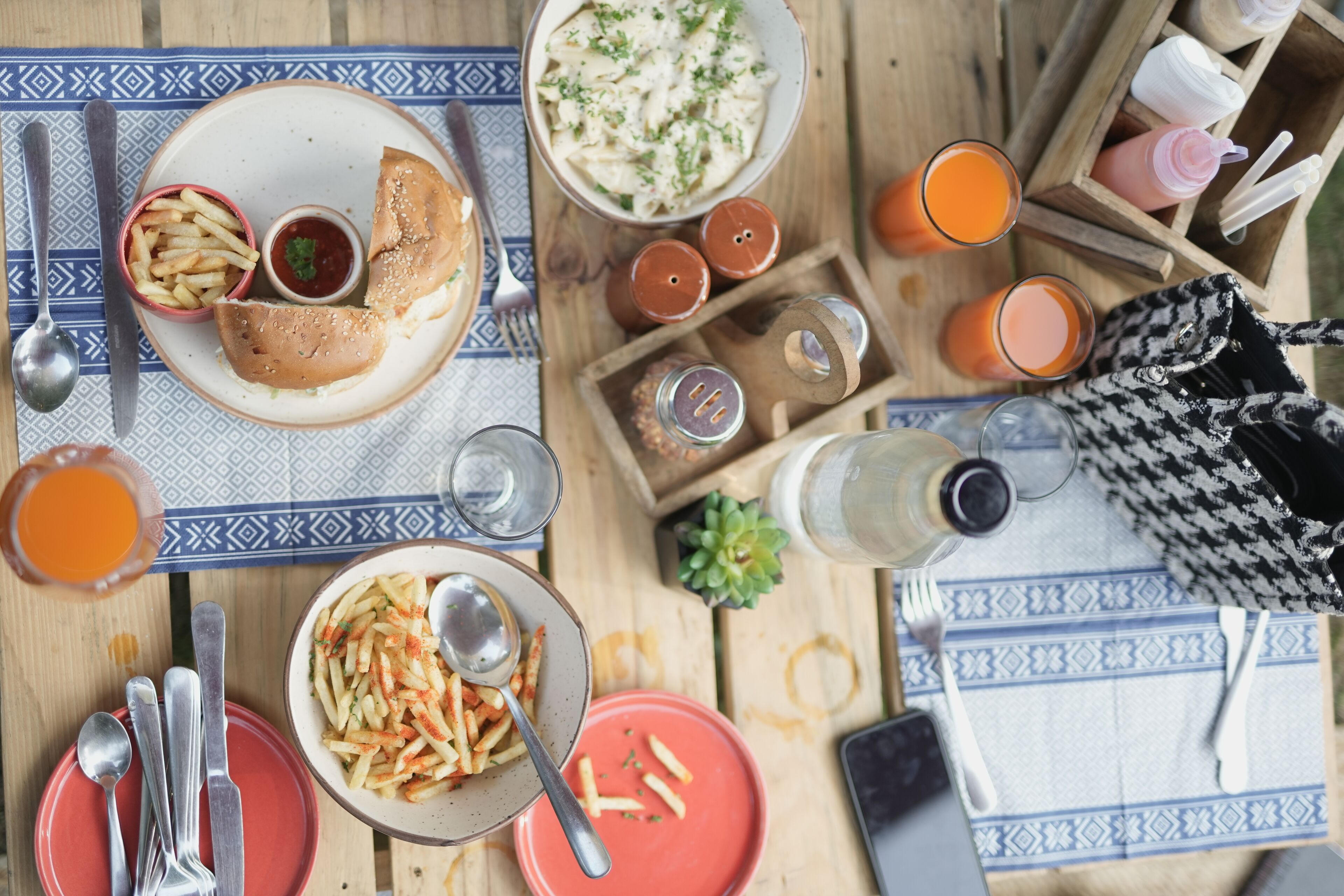 A table set with a meal, including a burger, fries, and pasta, along with various condiments and drinks.