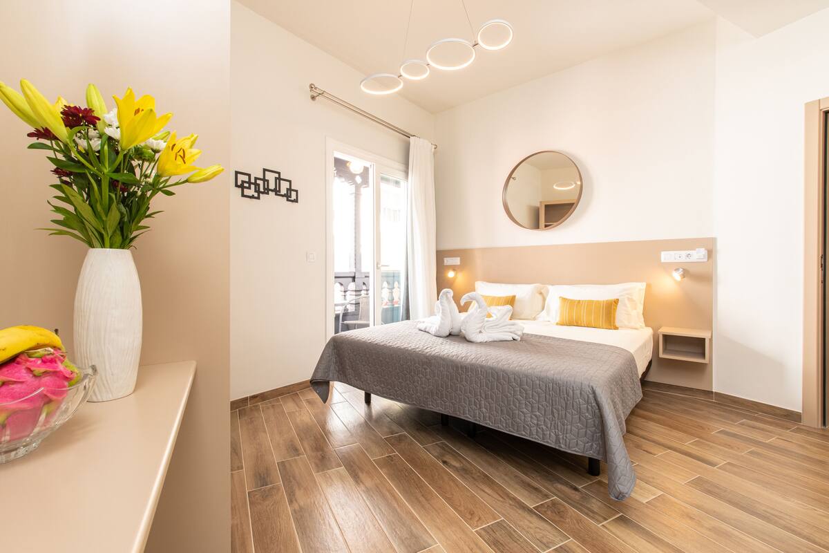 A modern hotel room with a bed, a bedside table, a mirror, and a vase of flowers.