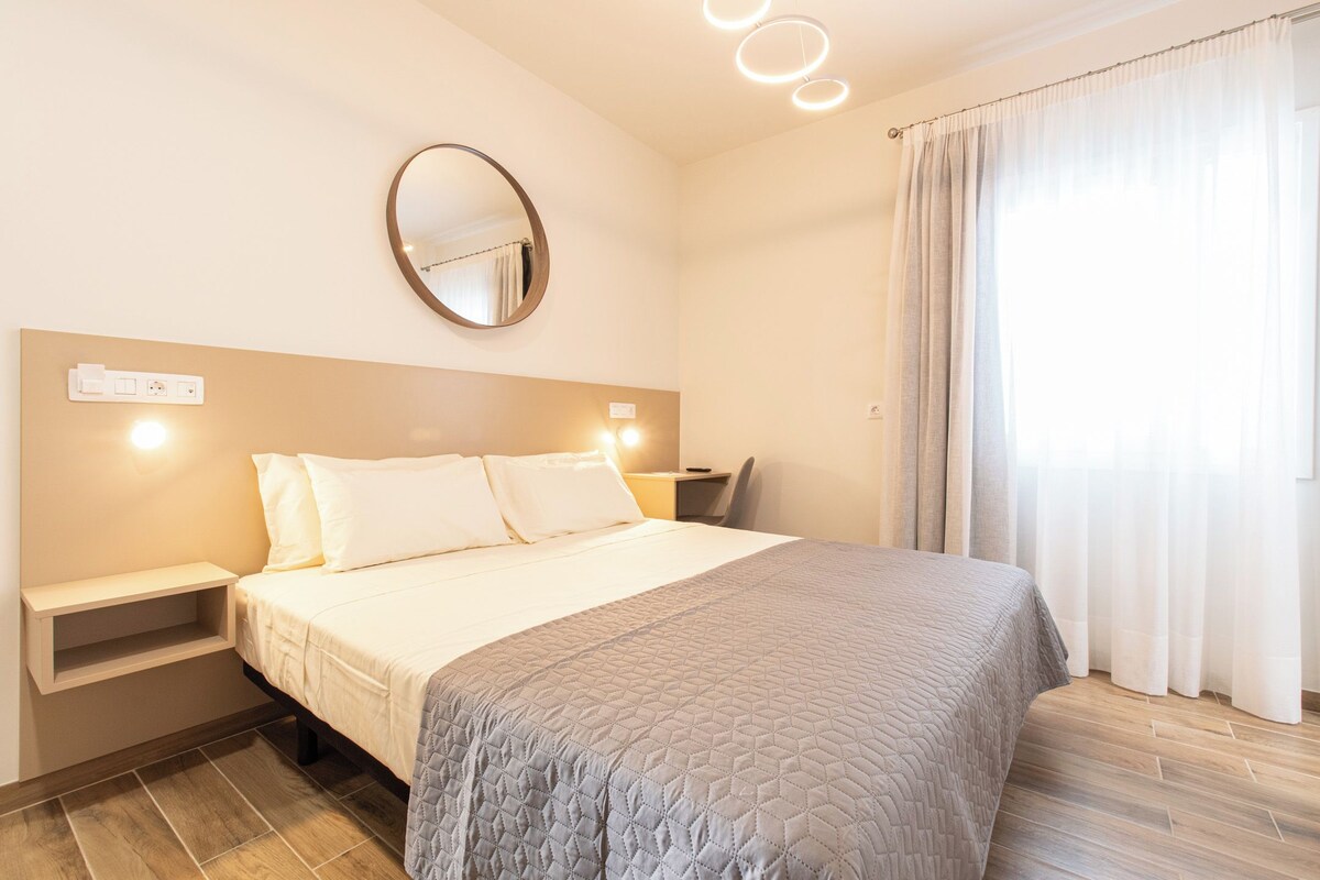 A hotel room with a bed, bedside table, wall-mounted mirror, and a window with curtains.
