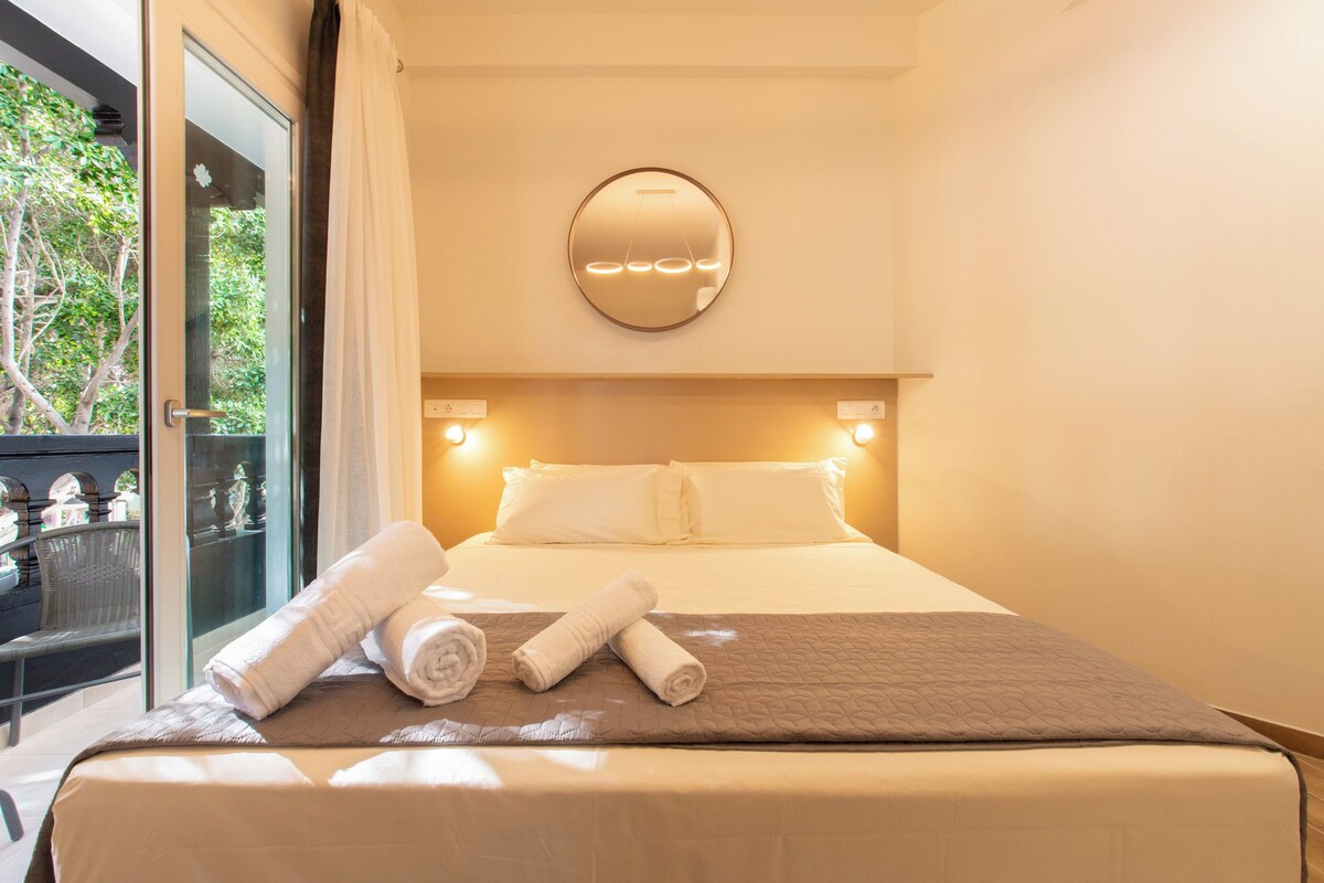A hotel room with a large bed, a round mirror on the wall, and a balcony with a view of trees.