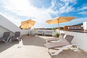 Eine Dachterrasse mit Liegestühlen, Sonnenschirmen, einem kleinen Tisch und Topfpflanzen.