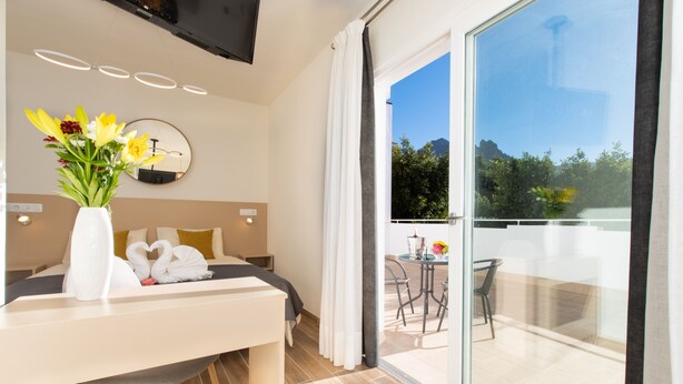 A modern hotel room with a bed, a desk with a vase of flowers, a round mirror, and a balcony with a table and chairs. - Adeje Plaza (Adeje)