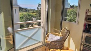Un balcón con una silla de mimbre, una estantería y vistas a zonas verdes y un edificio al otro lado de la calle.