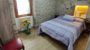 Un dormitorio con pared de piedra, techo de madera, cama con cubrecama estampado, una silla roja y una ventana con flores.