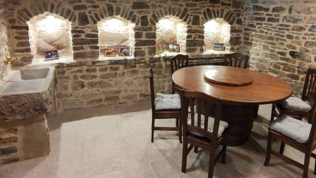 Habitación de paredes de piedra con una mesa redonda de madera y sillas, un lavabo de piedra y ventanas arqueadas con cuadros enmarcados.