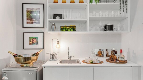A modern kitchen with a white countertop, a stainless steel sink, and a range hood. There are framed pictures on the wall, a wine rack, and a variety of glassware and bottles on the shelves. - CLUBHOUSE POKOLBIN - THE VINTAGE HOLIDAY HOUSES (POKOLBIN)