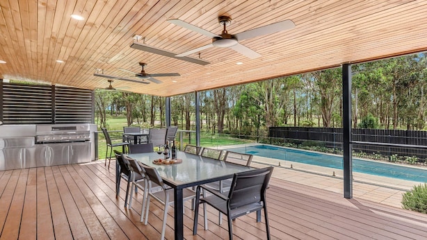 Un espacio al aire libre techado con piscina, terraza de madera, ventiladores de techo y una parrilla.