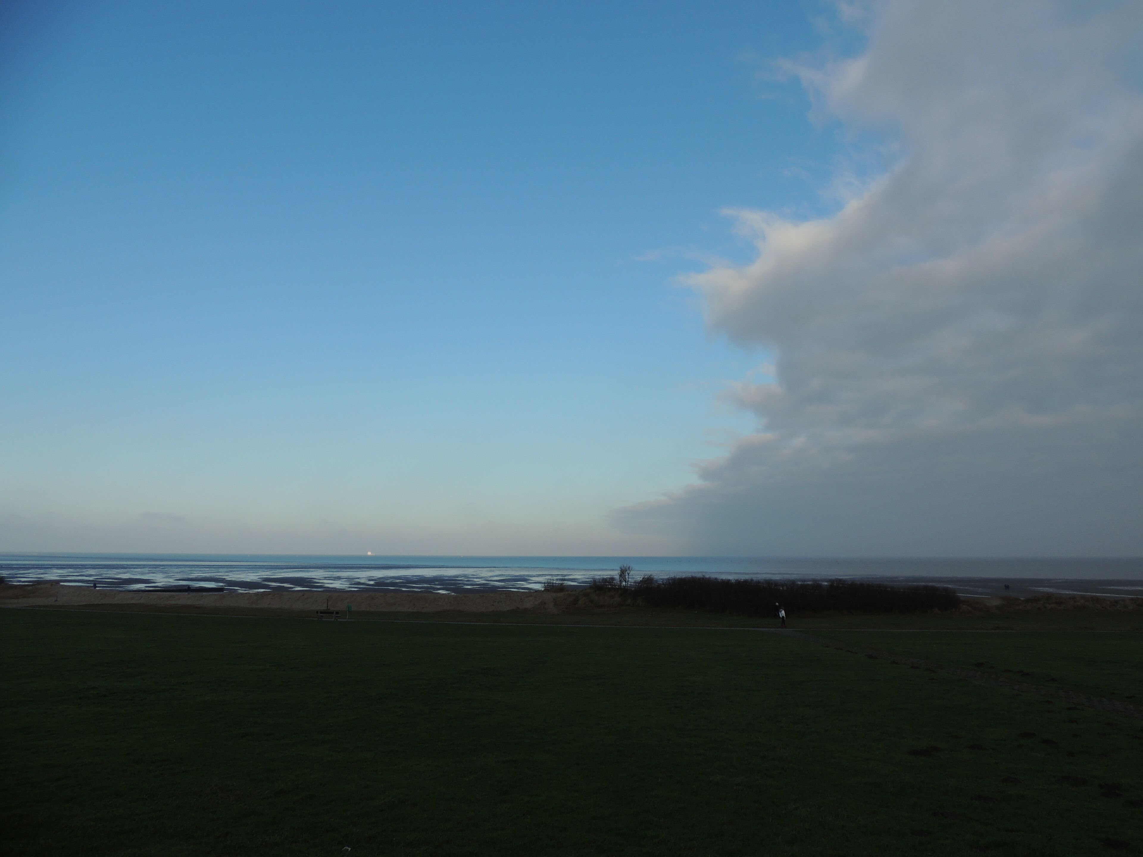 A coastal landscape with a clear sky, a grassy field, and a distant shoreline.
