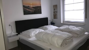Ein Schlafzimmer mit Bett, weißer Bettwäsche, einem Nachttisch mit Lampe, einem Fenster mit Jalousien und einem Bild an der Wand.