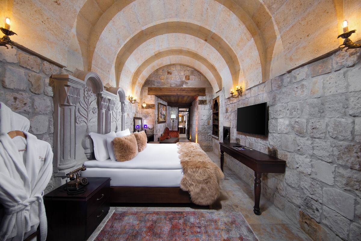 A bedroom with a stone wall, a bed with white bedding, a wooden nightstand, a robe, and a flat-screen TV.
