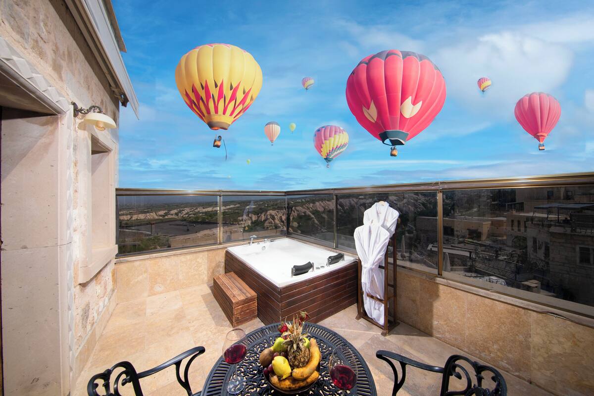 A balcony with a hot tub, a table with fruit and wine glasses, and a view of a cityscape.