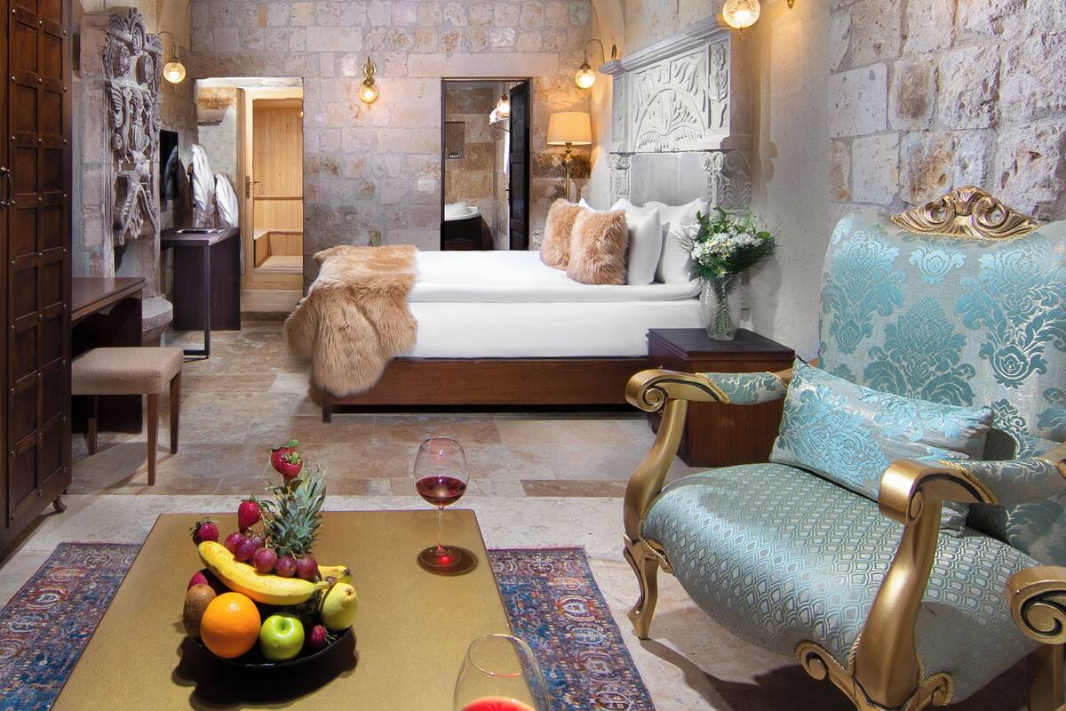 A hotel room with a large bed, a sitting area with a sofa, a dining table with fruit and wine, and stone walls.