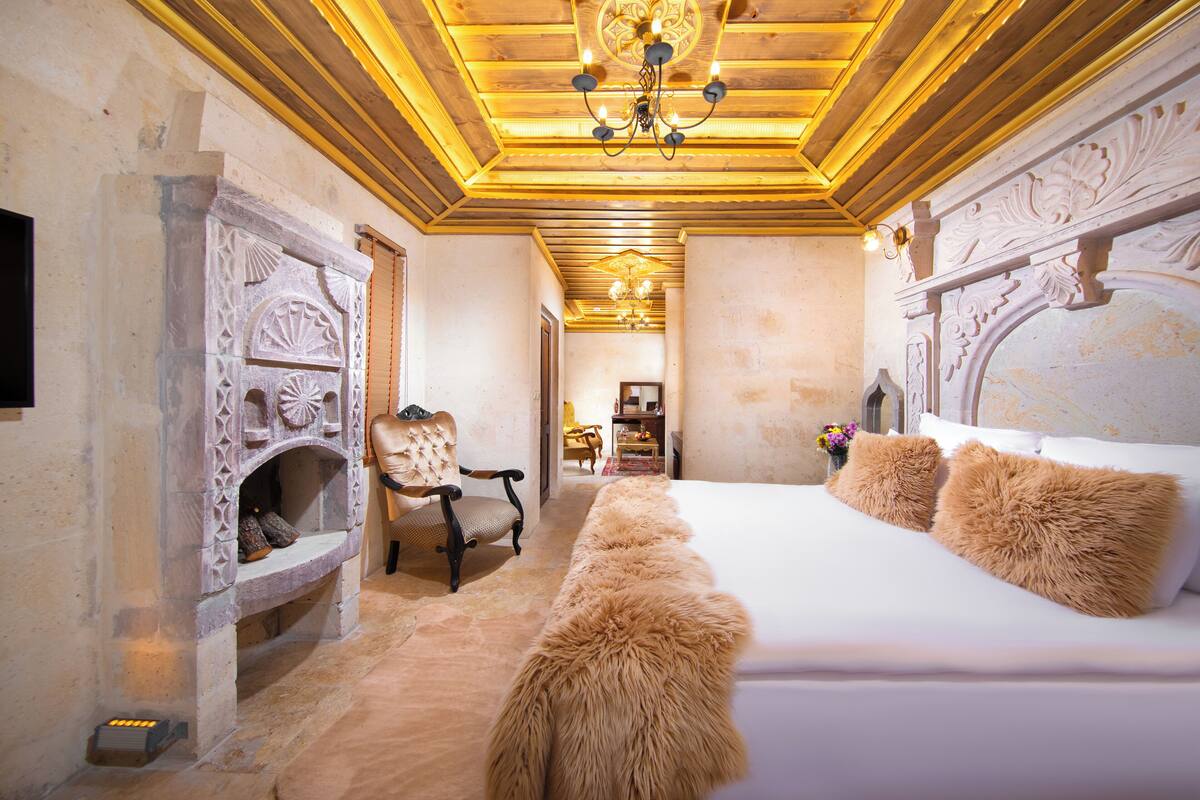 A bedroom with a large bed, a stone fireplace, a wooden ceiling, and a chandelier.