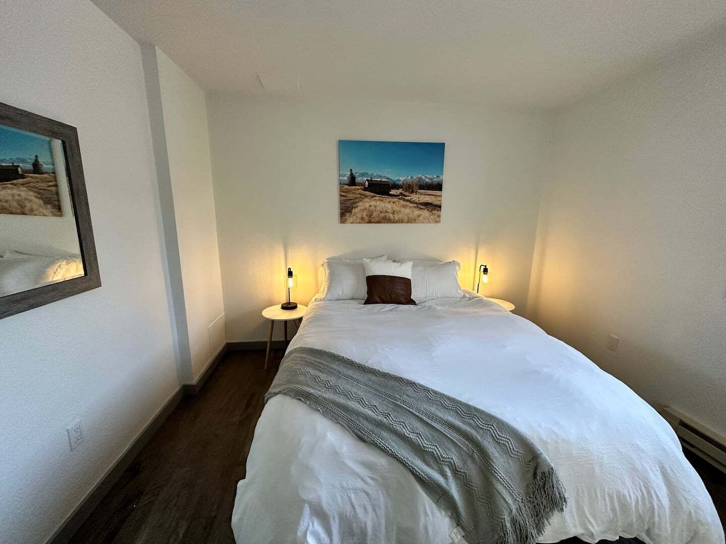 Cama bem arrumada com manta cinza, duas luminárias de cabeceira e uma pintura emoldurada de paisagem sobre a cama.