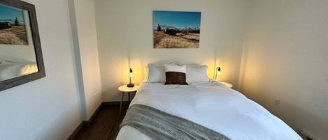 Cama bem arrumada com manta cinza, duas luminárias de cabeceira e uma pintura emoldurada de paisagem sobre a cama.