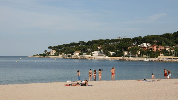 Een strand met mensen die zonnebaden en zwemmen, een heldere lucht en gebouwen op de achtergrond.