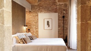 Een slaapkamer met een bakstenen muur, een bed met witte beddengoed, een lijstwerk kunstwerk aan de muur en een vloerlamp.