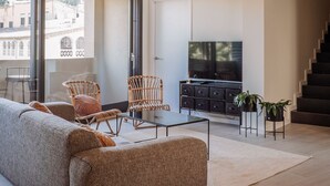 Een ruime woonkamer met een grote bank, rieten stoelen, een glazen salontafel en een televisie op een zwarte kast. Er hangen rieten hanglampen aan het plafond en een trap is op de achtergrond te zien.