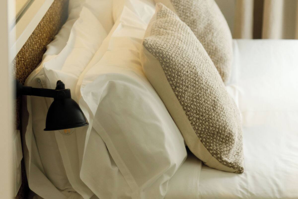 A neatly made bed with white linens and beige pillows, a black lamp on the side, and a mirror reflecting a potted plant.