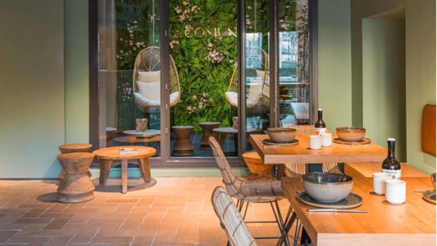 Een terras met tafels en stoelen, een hangstoel en een glazen deur met een groene wand, waarop de tekst “COM” zichtbaar is.