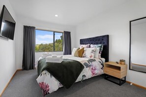 A bedroom with a bed, a bedside table, a TV, and a window with curtains. - Millar's Hideaway - Lake Views, Walk to Beach (Rotorua)