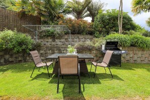 An outdoor patio with a table and chairs, a grill, and a stone wall with vegetation. - Millar's Hideaway - Lake Views, Walk to Beach (Rotorua)