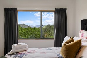 A hotel room with a bed, a window with curtains, and a view of greenery and mountains. - Millar's Hideaway - Lake Views, Walk to Beach (Rotorua)