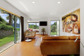 A spacious living room with a large window, a sofa, a chair, and a painting on the wall. - Millar's Hideaway - Lake Views, Walk to Beach (Rotorua)