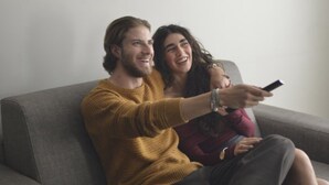 Un couple est assis sur un canapé gris dans une pièce où est accrochée, au mur, une peinture représentant un paysage.