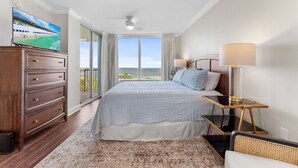 A bedroom with a large bed, a TV on a dresser, a lamp, and a view of the ocean.