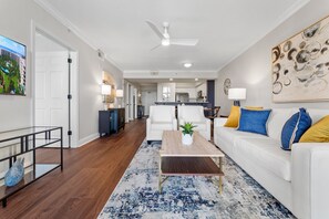 A modern living room with a white sofa, a coffee table, and a TV. There is a kitchen in the background with cabinets and a dining area. - Destin West Pelican 603  ~ Newly Remodeled ~ Lazy River! (Fort Walton Beach)