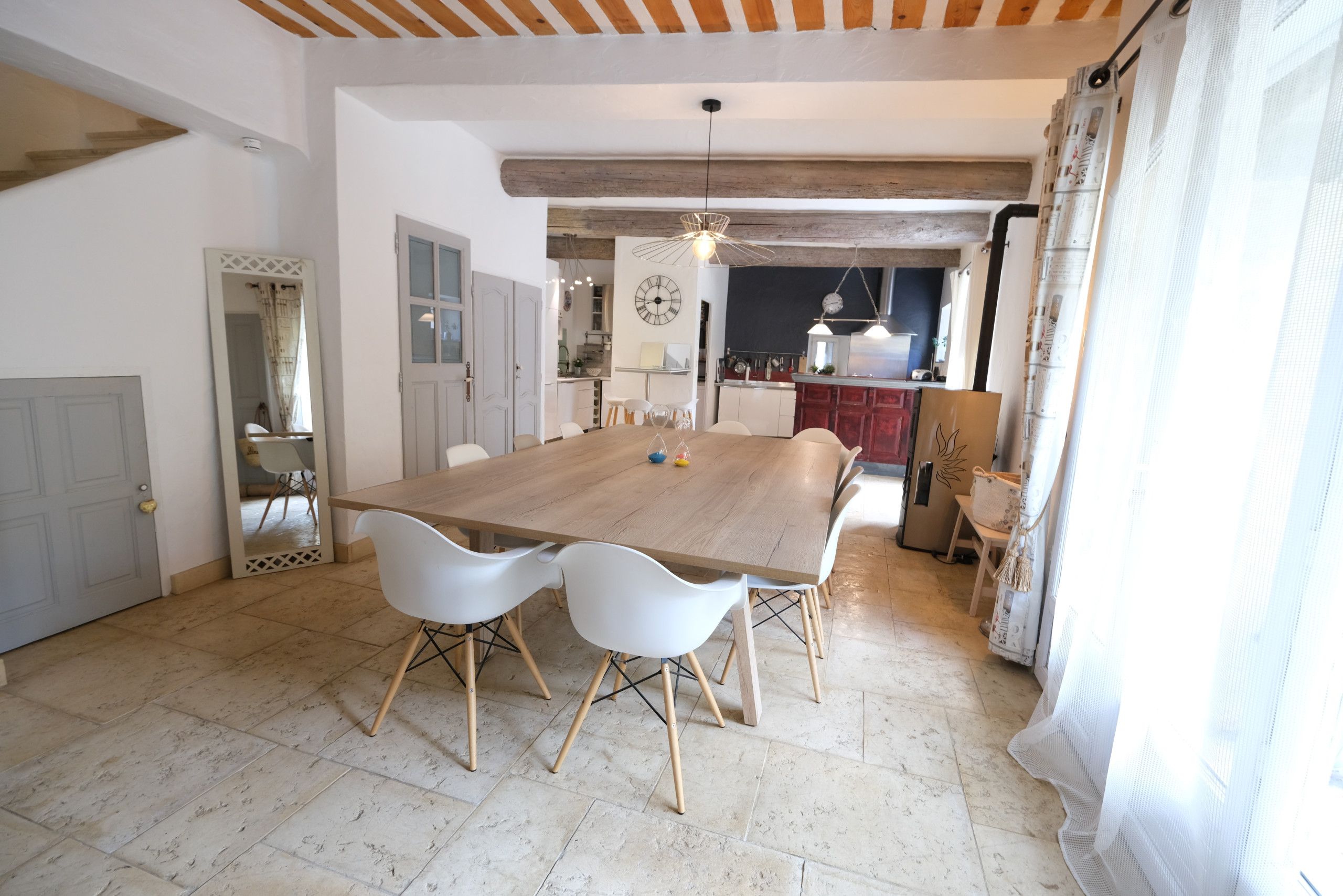 Un vaste espace de salle à manger avec une grande table en bois et des chaises blanches, une cuisine en arrière-plan et une vue sur l’extérieur par une fenêtre.