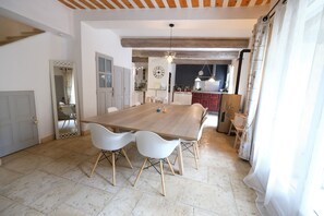 A spacious dining area with a large wooden table and white chairs, a kitchen in the background, and a view of the outdoors through a window. - Vacation rental with pool CHEVAL BLANC LS2-413 (Cheval Blanc)