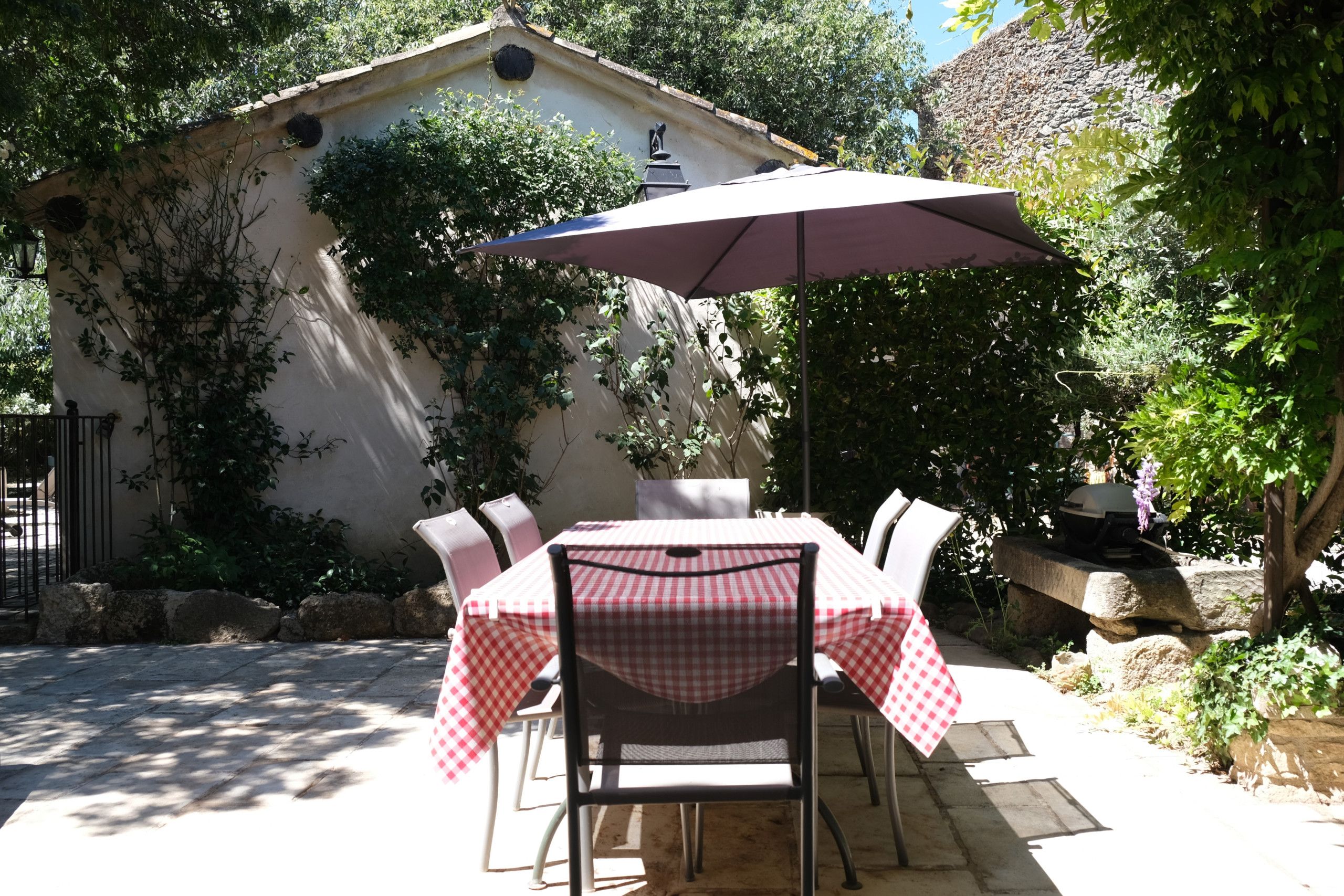 Une terrasse extérieure avec une table et des chaises recouvertes d’une nappe à carreaux, un grand parasol et un mur de pierre orné de plantes grimpantes.