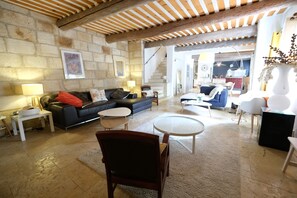 A spacious living area with a stone wall, a wooden ceiling, and a staircase leading to an upper floor. There are multiple seating options including a sofa, chairs, and a round table. - Vacation rental with pool CHEVAL BLANC LS2-413 (Cheval Blanc)