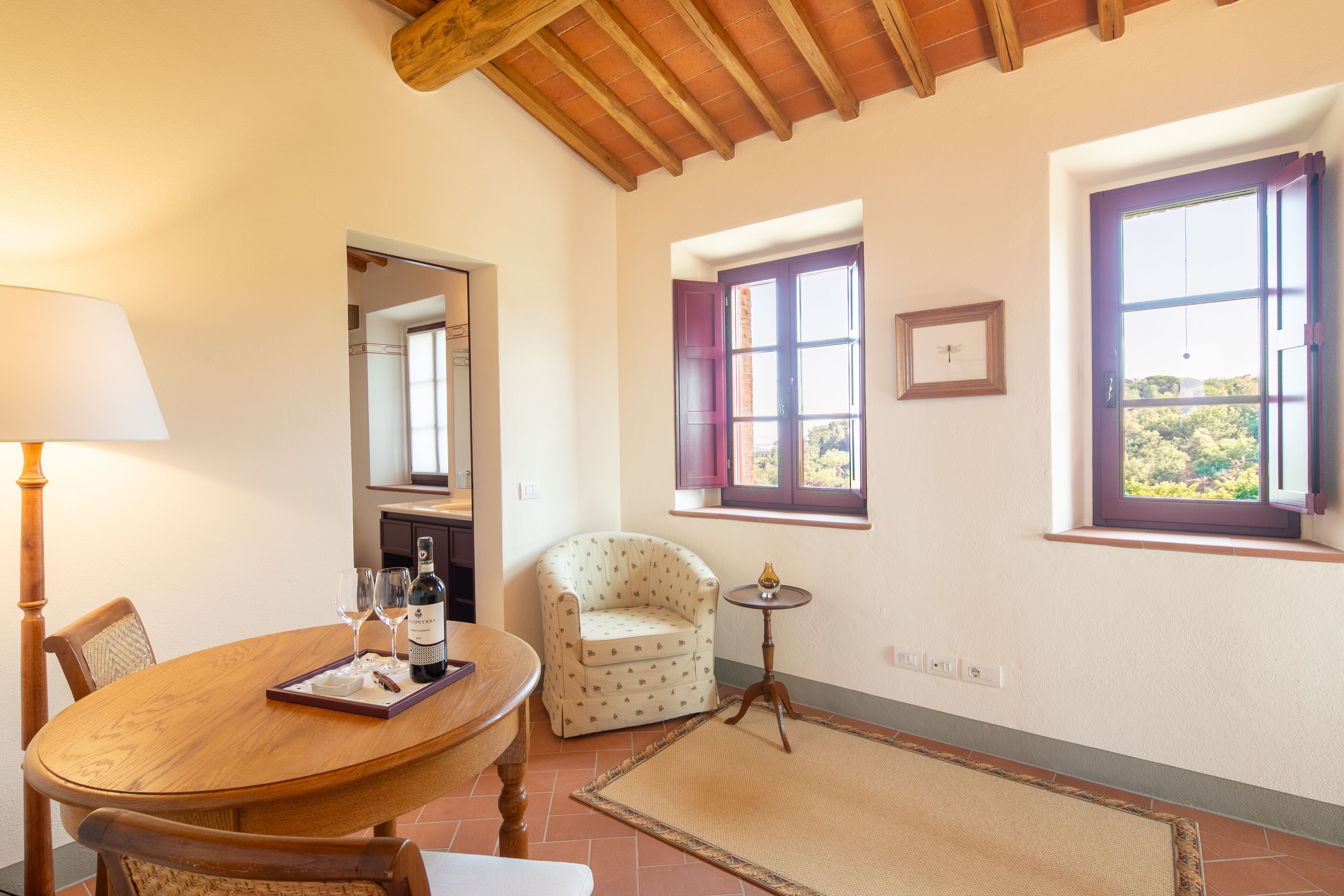 Un comedor con una mesa de madera, sillas, una lámpara, una pequeña mesa redonda con una botella y vasos, un sillón estampado de lunares, y una ventana con vistas a follaje.