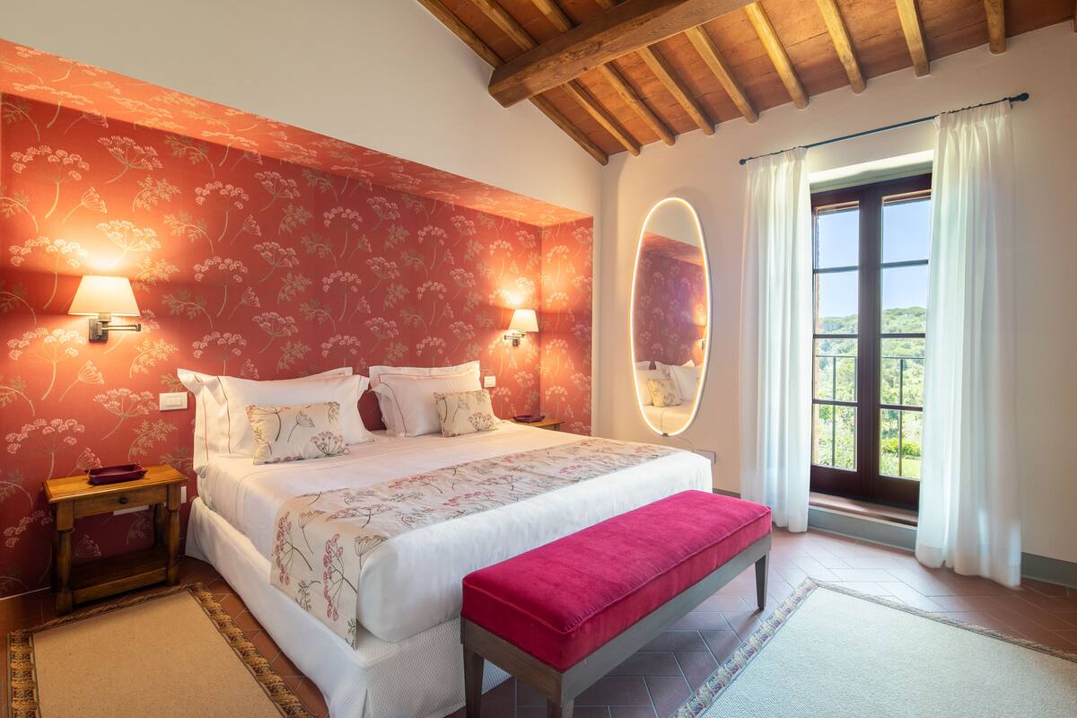 A bedroom with a large bed, two bedside lamps, a mirror, a bench, and a window with curtains.