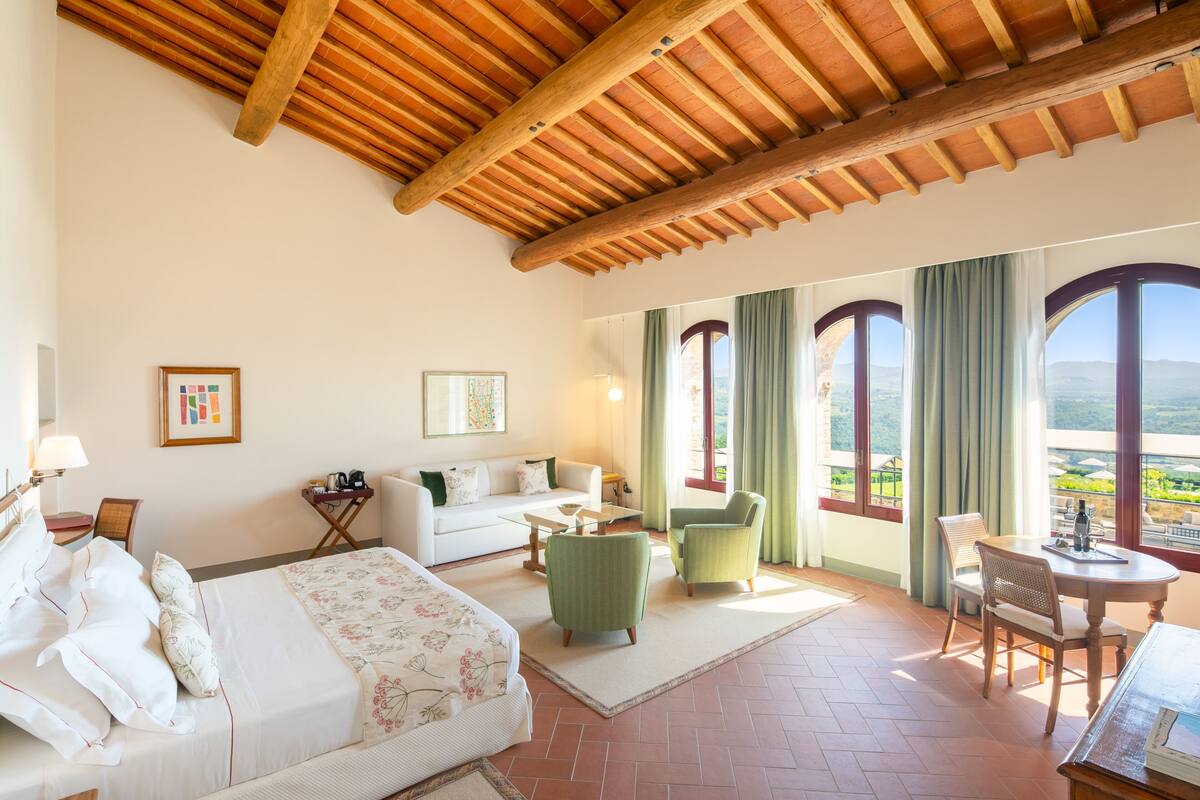 A hotel room with a bed, a sofa, two armchairs, a dining table, and large windows with curtains.
