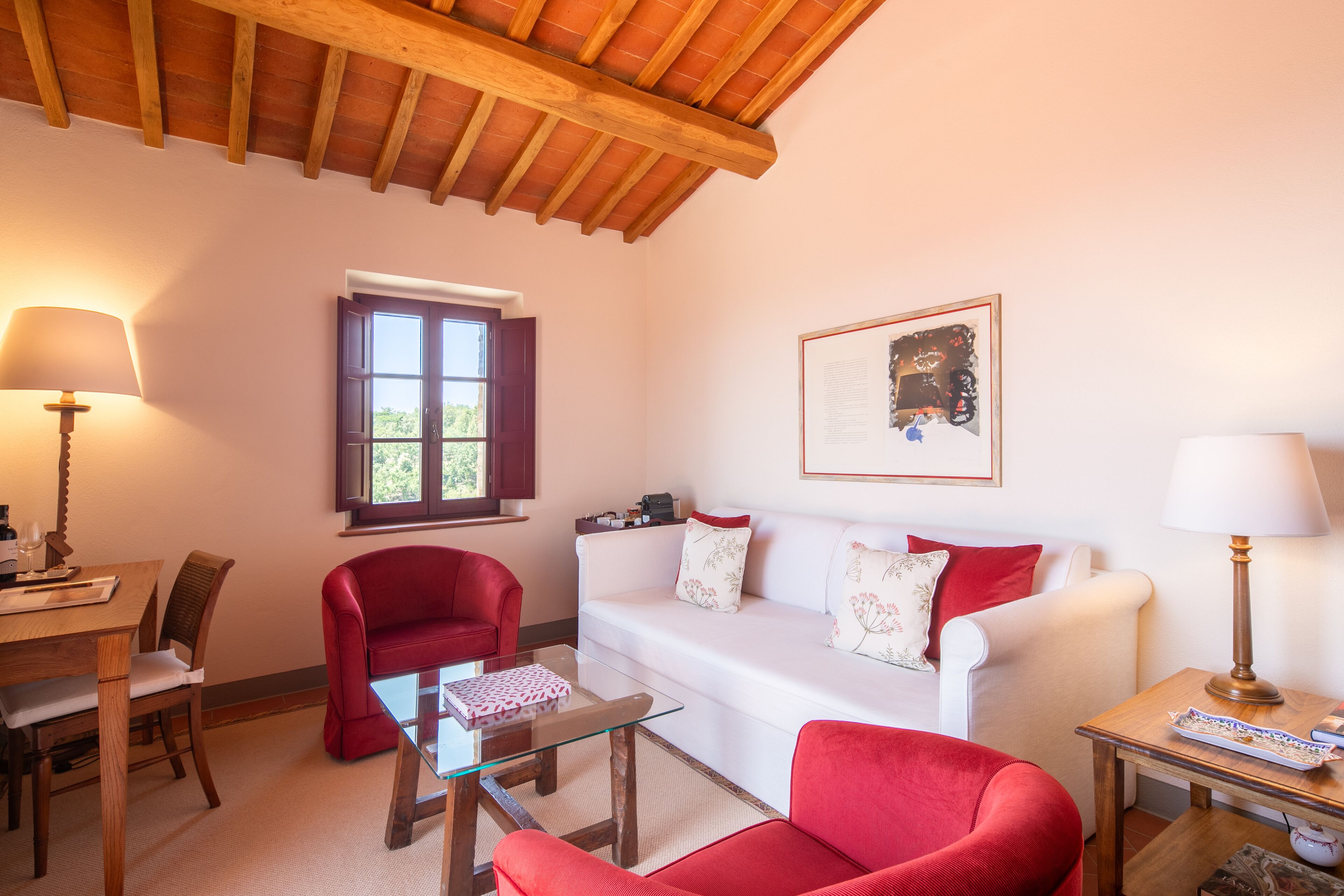 Un soggiorno con soffitto in legno, un divano bianco con cuscini rossi, un tavolino da caffè in vetro, una poltrona rossa, un tavolino laterale in legno con una lampada e una finestra con vista sul verde.