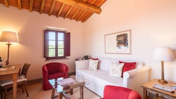 Un soggiorno con soffitto in legno, un divano bianco con cuscini rossi, un tavolino da caffè in vetro, una poltrona rossa, un tavolino laterale in legno con una lampada e una finestra con vista sul verde.