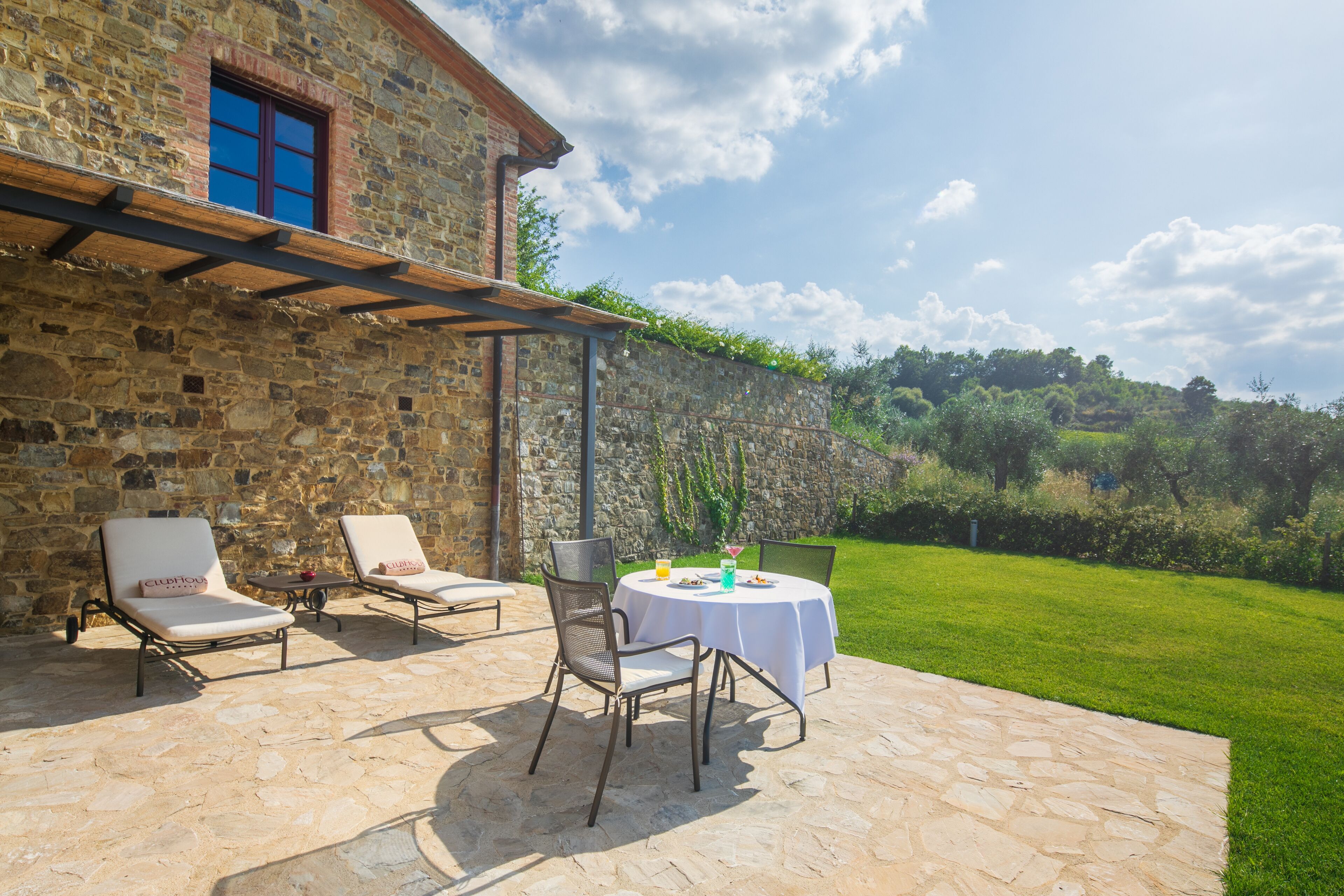 An outdoor patio with stone flooring, two lounge chairs, a table with a white tablecloth, and a stone wall.
