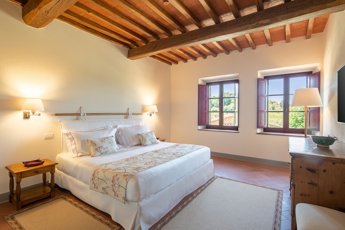 A bedroom with a large bed, wooden headboard, two bedside lamps, a dresser, and two windows with shutters.