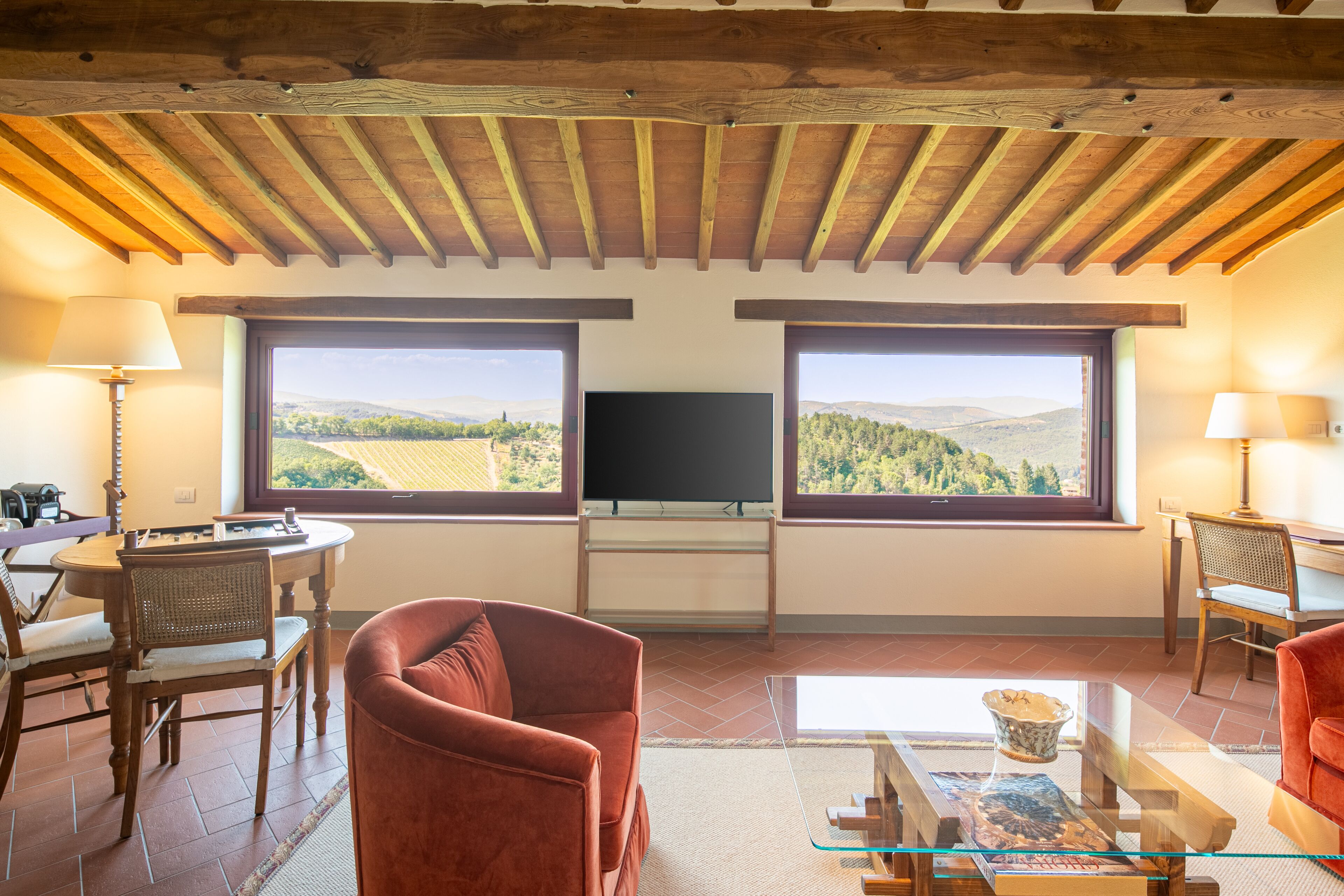 A living room with a TV, two large windows with a view, a red sofa, and a glass coffee table.